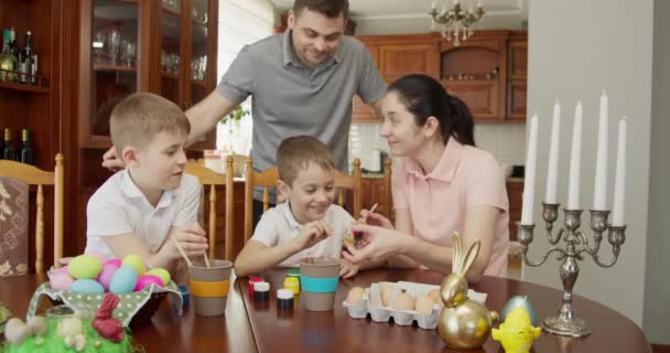 Az apa hátulról öleli meg a feleségét és a két gyerekét. A család húsvéti tojásokat fest. — Stock videók