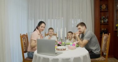 İki genç çocuğun ebeveynleri bilgisayar kullanarak video konuşuyorlar, çevrimiçi televizyon yayınlarına göz atıyorlar, hafta sonları evde, Paskalya için hazırlanan tatil masasında birlikte vakit geçiriyorlar.