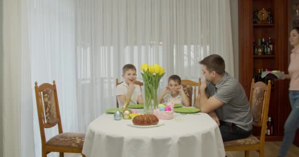 Alegre madre trae una cesta de huevos pintados y lo pone sobre la mesa mientras sirve la cena de vacaciones para una familia numerosa — Vídeos de Stock