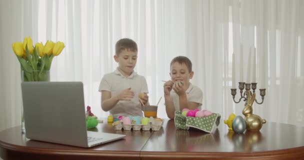 Dois rapazes pintam ovos de Páscoa e cumprimentam-se. sobre a mesa é um computador que os meninos estão assistindo e ter uma videoconferência — Vídeo de Stock