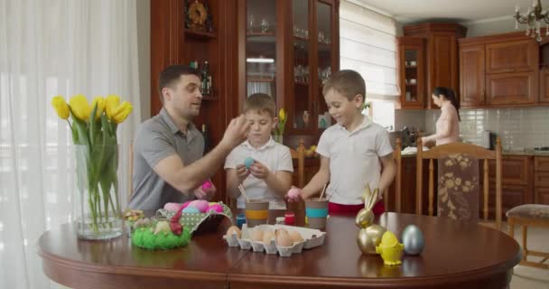 Vater mit zwei Jungen jongliert mit bunten Eiern, meine Mutter erledigt Hausarbeiten in der Küche. Osterstimmung — Stockvideo
