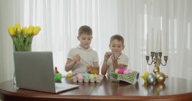 İki çocuk bilgisayara bakarak yumurtalarını boyuyor.
