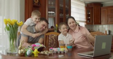 Anne, baba ve iki çocuk bir masada oturup bilgisayar kullanarak görüntülü konuşma yapıyorlar. Bir çocuk babasının sırtına oturur ve onu kucaklar.