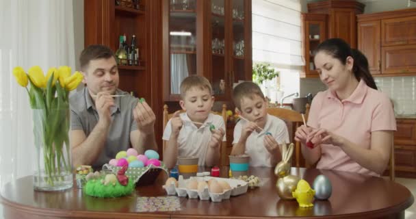 Eine glückliche Familie sitzt an einem Tisch und bemalt Ostereier. sie alle strecken die Hand aus und zeigen bunte Eier — Stockvideo