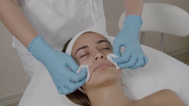 Em uma clínica de beleza, o cosmetologista limpa a pele do rosto de uma jovem cliente feminina com um cotonete, a mulher está deitada na mesa com os olhos fechados — Vídeo de Stock