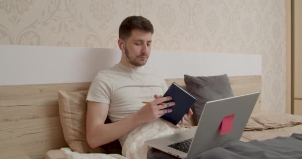 El chico está trabajando en la computadora en la cama. tiene una tarjeta azul en la mano. leer del cuaderno en una videollamada al ordenador — Vídeo de stock