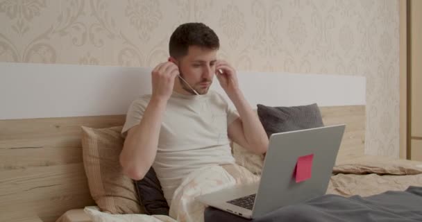 Chłopiec w białej koszulce leżący w łóżku. Zakłada słuchawki i patrzy na komputer. — Wideo stockowe
