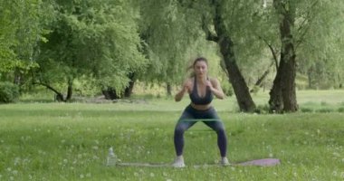 Kahverengi saçlı çekici bir kadın, kalçasını ve bacak kaslarını doğadaki çimlerin üzerinde, mor bir halıda eğitiyor..