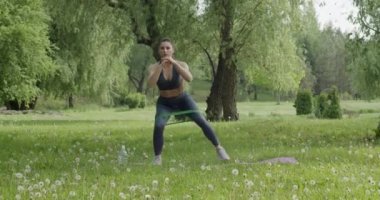 Kahverengi saçlı çekici bir kadın, kalçasını ve bacak kaslarını doğadaki çimlerin üzerinde, mor bir halıda eğitiyor..