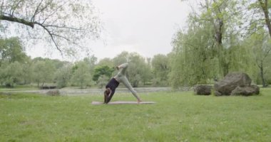 Yoga yapan genç bir kadın, geniş açılı formda bir kadın diz kaldırma pozu veriyor yeşil bir bahçede pratik yapıyor..