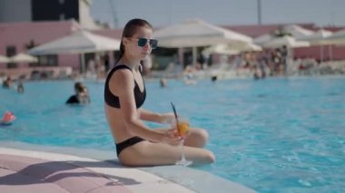 girl at a tropical pool sunbathing on the edge of the pool, drinking cocktail. relaxing on a summer day at the pool