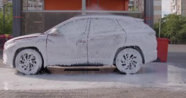 A man washes his car during the day, washes of the foam. A red SUV car at a self-service car wash. Washing the car with your own hands. A red auto in cleaning foam.