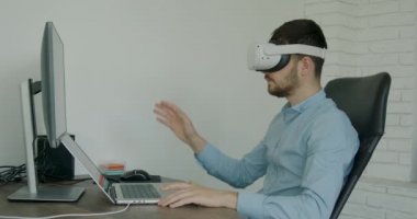 Man in blue shirt sitting at desk and using VR glasses and laptop for work. A freelancer swiping the virtual screen to the right with his fingers.