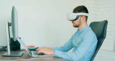 young man with VR glasses typing on the computer in a bright office. man in a blue shirt in a bright office typing on a computer with VR glasses. vr concept