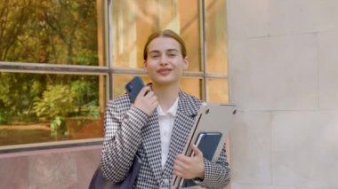 Portrait of young brunette wearing braces. Female person standing in front of a building and smiling widely. Close-up with a business woman holding a computer, phone and a notebook.