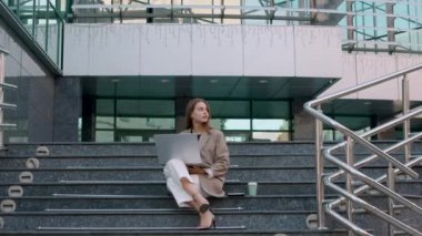 confident young business woman in suit and heels, working on laptop and drinking coffee from plastic cup, standing on the stairs of a business center. Free job and business woman concept 