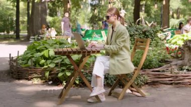 independent woman sitting at a table on the summer terrace of a cafe, classically dressed and wearing sunglasses, typing on a computer. Sunny day. The concept of freelancer
