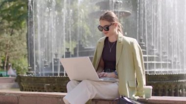 Elegant young woman sitting on the fountain in the city center, working on the computer. Freelancer using computer while resting in the city on a sunny summer day. business woman concept