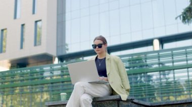 Close-up portrait of a young woman sitting on the stairs outside the business center typing on her laptop and surfing online. He looks at the clock to see how much time he has left. Business concept