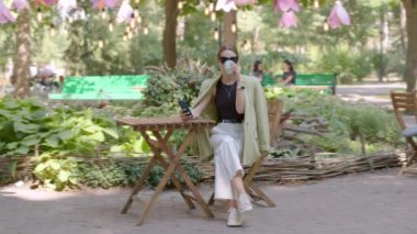 Cheerful young woman wearing stylish outfit and sunglasses sitting in the city at a terrace, using a new modern smartphone and drinking coffee. Communication, online shopping, social network