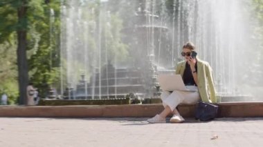business woman is talking on the phone and at the same time working on the computer. Sitting down by a fountain, with sunglasses and a coffee. concept of independent, business and successful woman