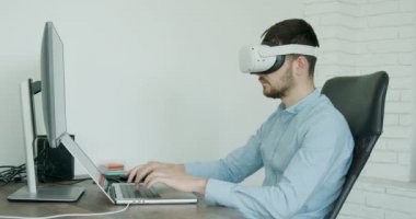  Mavi gömlekli bir adam parlak bir ofiste sanal gerçeklik gözlüklü bir bilgisayarda yazı yazıyor. Gözlüklü genç adam parlak bir ofiste bilgisayarda daktilo kullanıyor. vr kavramı