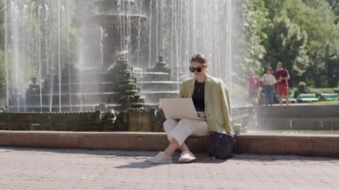 Uygun bir işle istediği yerden çalışan bir kadının uzak bir görüntüsü. Görevlerini parkta çözen bir bayan yönetici, bir göletin yanında oturuyor.