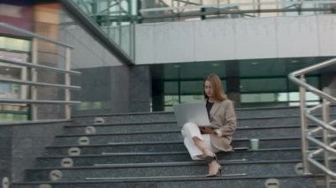 Klasik ve topuklu giymiş modern bir ofisin merdivenlerinde oturup bilgisayar başında çalışan bir kadının etkileyici görüntüsü. Her yerde çalışan kadın yönetmen.