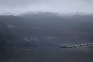Ağaçların arasından Norveç göl nehrinin bulutlu ve yağmurlu havası görünüyor. Kasım 'da Norveç. Nehir gölünün yanında dinlenme yeri. Norveç 'te dağ manzarası.