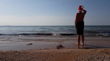 Yüzme şortlu bir genç gün batımında kumlu sahilde Noel Baba şapkasıyla duruyor. Kumsalda Noel