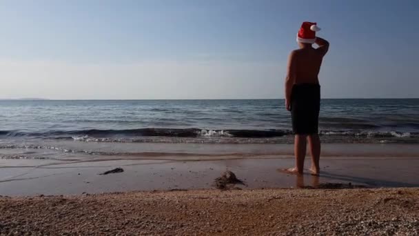 Nastolatek w kąpielówkach stoi na piaszczystej plaży nad morzem o zachodzie słońca w kapeluszu Świętego Mikołaja. Boże Narodzenie na plaży — Wideo stockowe