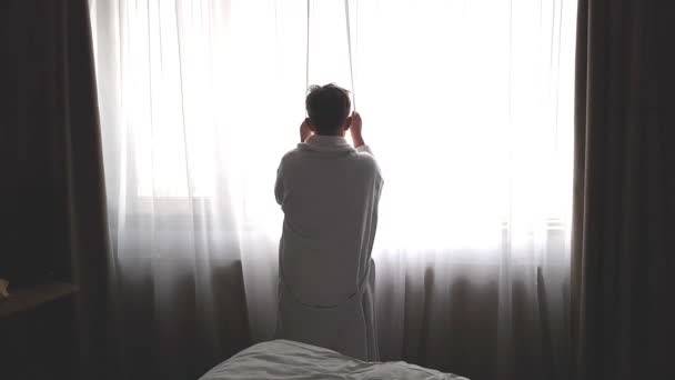 Un adolescente con un albornoz blanco se para en la ventana y abre las cortinas. mañana en el hotel — Vídeo de stock