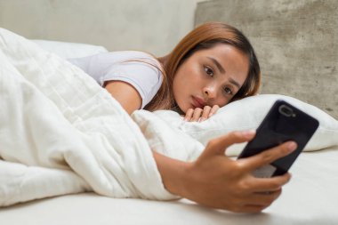 mutsuz Asyalı güzel kız yatakta akıllı telefon kullanarak üzgün hissediyor.
