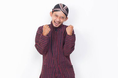 asian man with javanese traditional cloth lurik raised his arm happy and excited celebrating victory expressing big success on white background