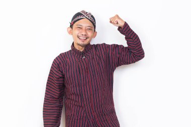 asian man with javanese traditional cloth lurik raised his arm happy and excited celebrating victory expressing big success on white background