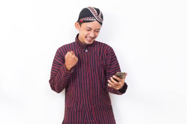 Happy joyful asian man with javanese traditional cloth lurik using mobile smartphone isolated on white background