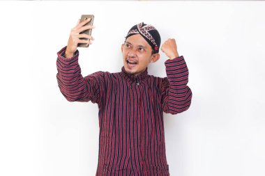 Happy joyful asian man with javanese traditional cloth lurik using mobile smartphone isolated on white background