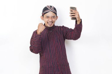 Happy joyful asian man with javanese traditional cloth lurik using mobile smartphone isolated on white background