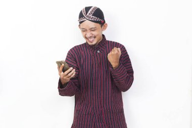 Happy joyful asian man with javanese traditional cloth lurik using mobile smartphone isolated on white background