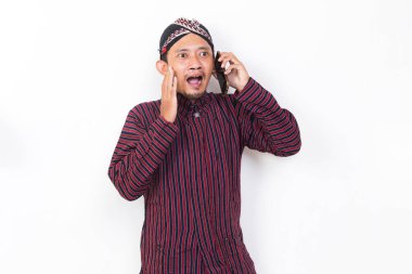 Happy joyful asian man with javanese traditional cloth lurik using mobile smartphone isolated on white background