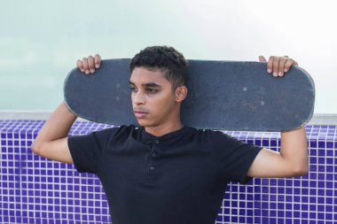 Genç Latin bir adamın kaykayla poz verirken yakın bir fotoğrafı. Düşünceli bir duruşu var..