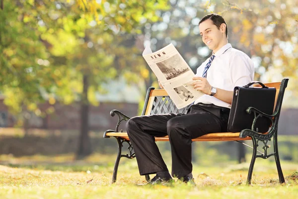 Молодой человек сидит на скамейке и читает газету в солнечный день. — стоковое фото