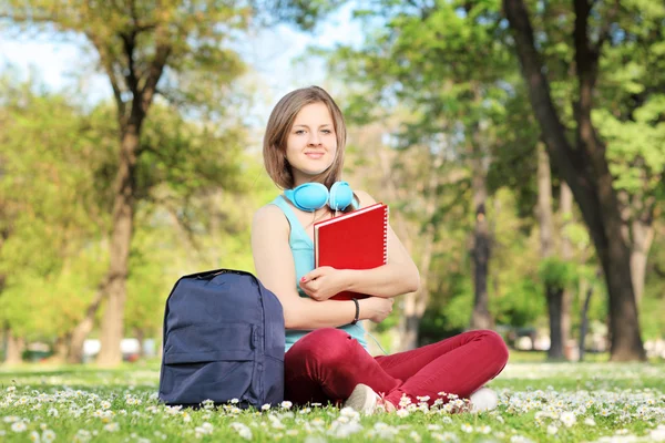 Estudante bonita com livro e fones de ouvido sentado em uma grama em um parque — Fotografia de Stock