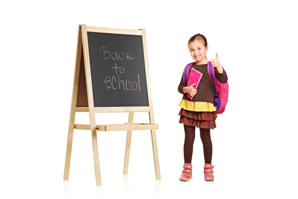 Child next to a school board holding book and showing thumb up