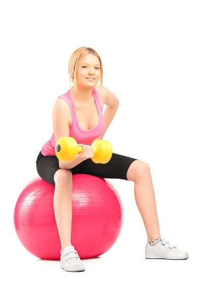 Blond female bodybuilder lifting up a dumbbell seated on a ball — Stock Photo, Image