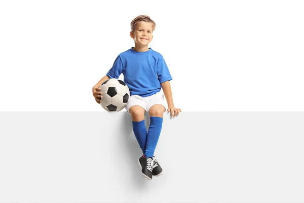 一个穿着蓝色足球球衣的可爱小男孩坐在一块空白的板子上 手里拿着一个被白色背景隔开的球 — 图库照片