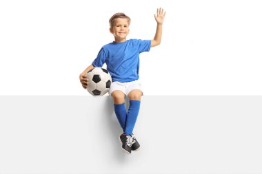 Beyaz arka planda izole edilmiş bir futbol topuyla boş bir panele el sallayan ve oturan çocuk