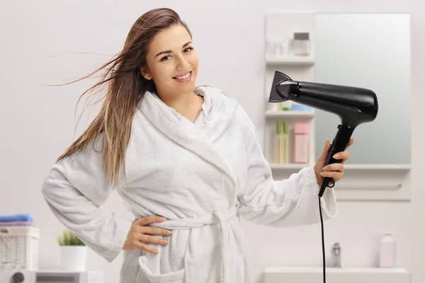 年轻女人穿着浴衣 在浴室里用吹风机擦干头发 — 图库照片