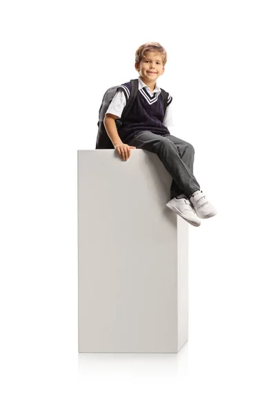 制服を着た男子生徒が白い柱の上に座り 白い背景に隔離されたカメラを見る — ストック写真