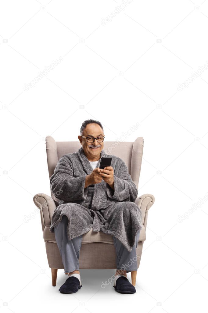 Mature man in a bathrobe sitting in an armchair and using a smartphone isolated on white background